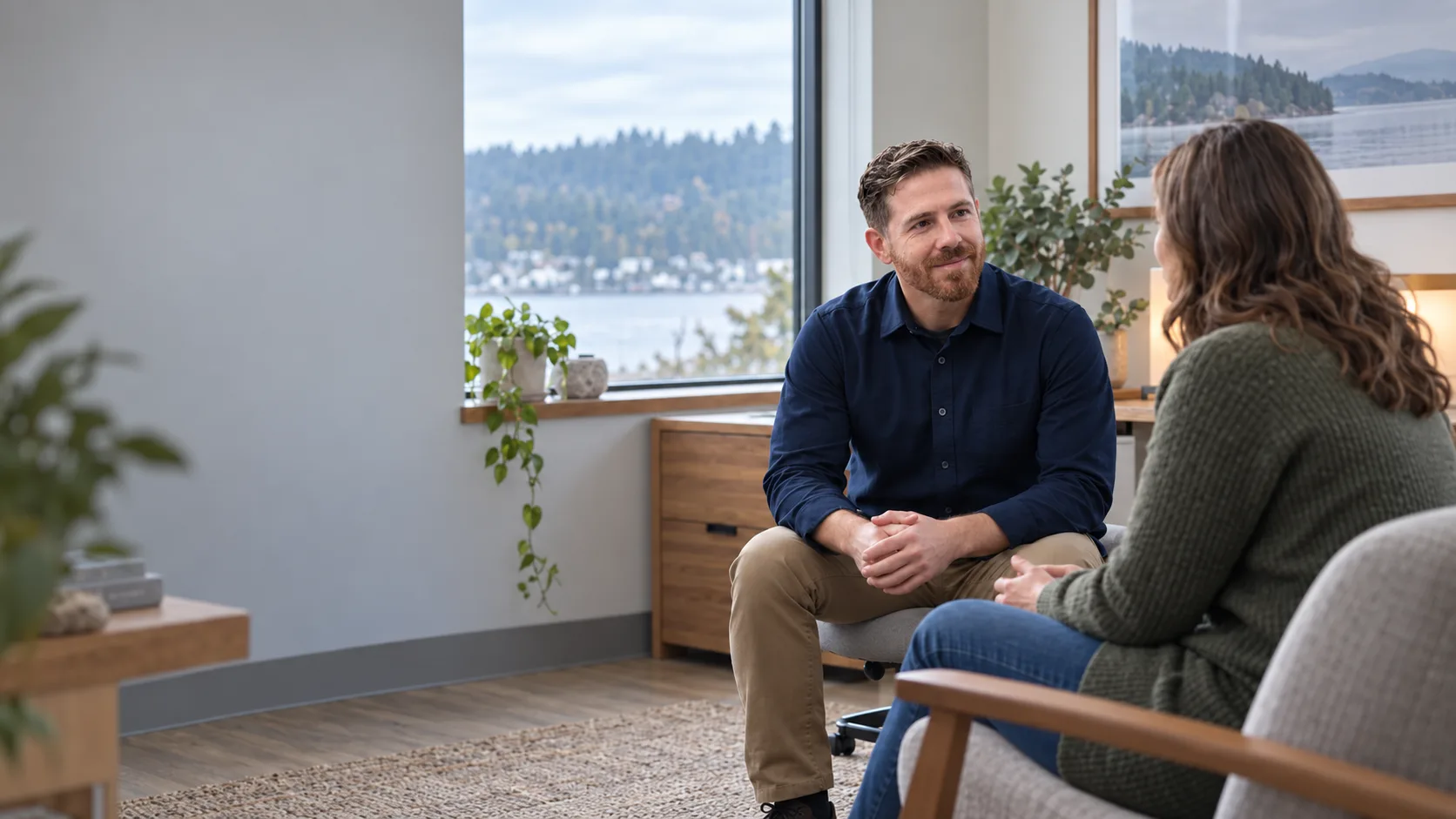 Dr. Mark Hilmy in a navy shirt seated and listening to an adult patient in a calm consultation room with a waterfront view