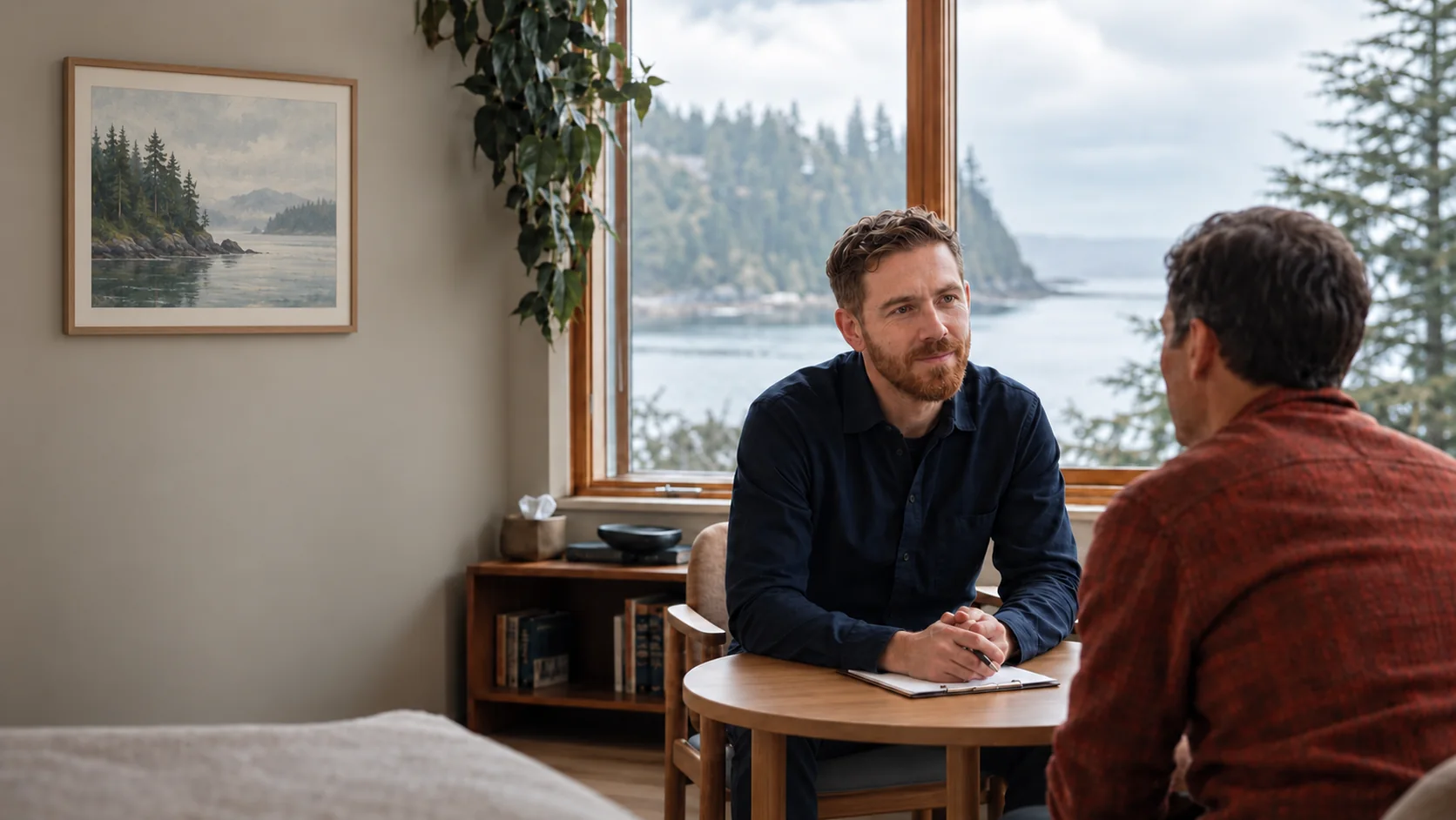 Dr. Mark Hilmy in a navy shirt listening to an adult patient across a round wooden table, with the waterfront visible through the window and a framed landscape on the wall